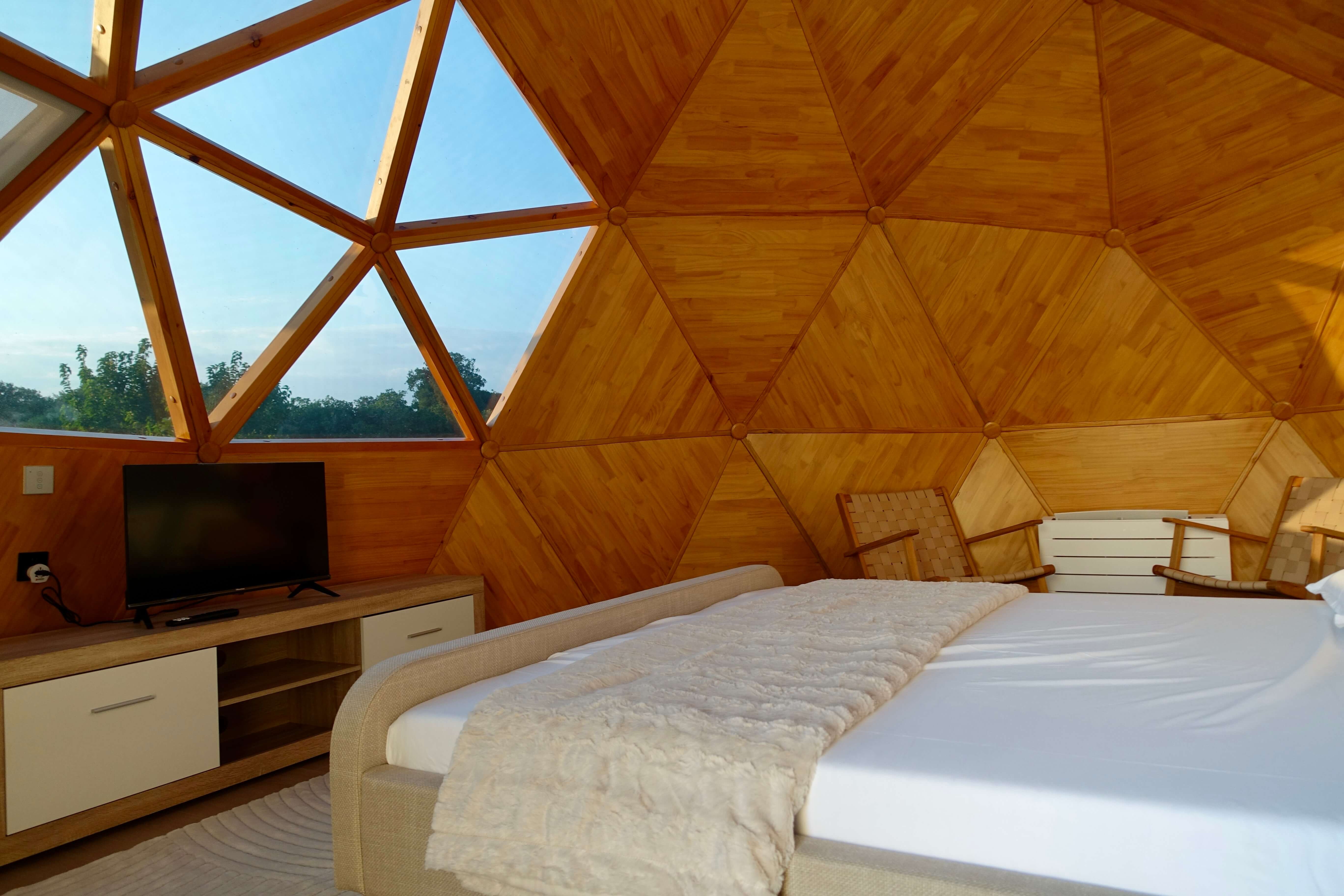 Bed, TV and glazed dome wall looking out to trees and open sky at Solar Dome Retreat